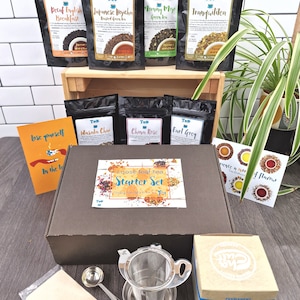 May include: A brown box containing a selection of loose leaf tea bags, a tea strainer, a tea scoop, and a card that reads "Loose leaf tea Starter Set". The box is placed on a wooden surface with a plant in the background.