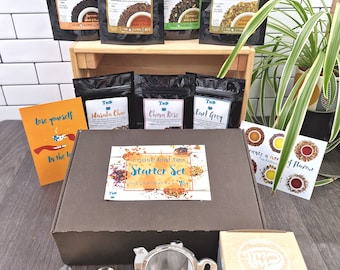 May include: A brown box with a variety of loose leaf tea bags, a tea strainer, a tea scoop, and a card that says "Loose leaf tea Starter Set". The box is sitting on a wooden surface with a plant in the background.