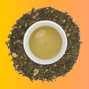 May include: A white ceramic cup filled with light green tea, surrounded by loose green tea leaves with bits of dried fruit and herbs.