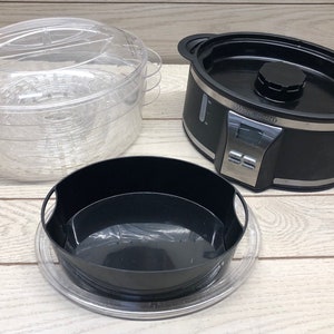 Puede incluir: Un vaporizador eléctrico negro con temporizador digital y una cesta de vapor de plástico transparente. El vaporizador tiene una base negra con un borde plateado y una tapa de plástico transparente.