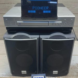 Pode incluir: Um sistema estéreo Pioneer com leitor de CD, duas colunas e um controlo remoto. O sistema tem um acabamento prateado com um visor azul que diz "PIONEER". As colunas são pretas e retangulares.