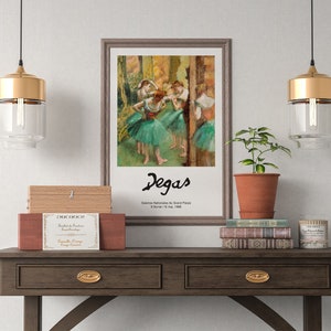 May include: A framed painting of dancers by Edgar Degas, titled "Classe de Danse" with the text "Galeries Nationales du Grand Palais, 8 November 1988 - 11 March 1989" below the signature. The painting is hanging on a white wall above a wooden table with two drawers, a stack of books, a potted plant, and two gold pendant lights.