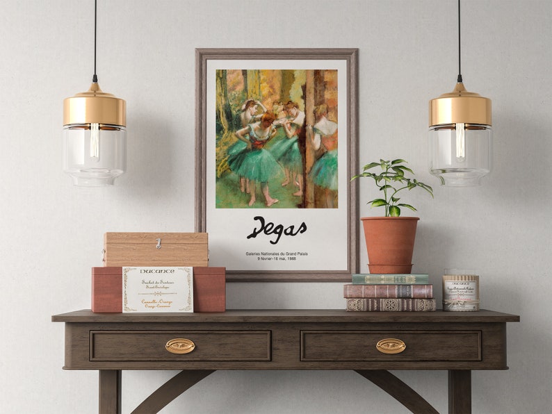 May include: A framed painting of dancers by Edgar Degas, titled "Classe de Danse" with the text "Galeries Nationales du Grand Palais, 8 November 1988 - 11 March 1989" below the signature. The painting is hanging on a white wall above a wooden table with two drawers, a stack of books, a potted plant, and two gold pendant lights.