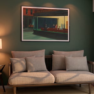 May include: A living room scene featuring a framed print of Edward Hopper's "Nighthawks" painting. The artwork hangs above a light gray sofa with matching pillows. A lamp with a white shade and a small side table with decorative plants complete the decor.