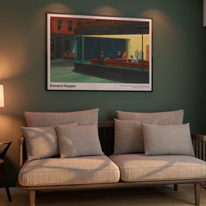 May include: A beige sofa with multiple pillows sits in a room with green walls and a framed print of Edward Hopper's "Nighthawks" hanging above it. A lamp with a white shade and a dark brown base sits on a side table next to a potted plant.