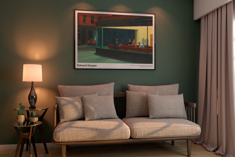 May include: A beige sofa with multiple pillows sits in a room with green walls and a framed print of Edward Hopper's "Nighthawks" hanging above it. A lamp with a white shade and a dark brown base sits on a side table next to a potted plant.