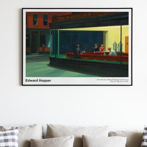 May include: A painting by Edward Hopper titled "Nighthawks" depicting a diner at night with three customers and a server. The diner is brightly lit with a yellow glow, while the street outside is dark. The painting is framed in black.