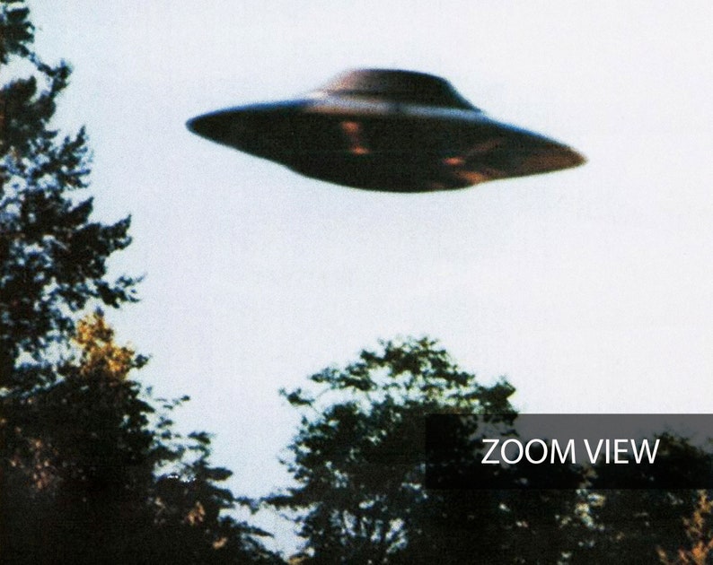 May include: A black flying saucer shaped object against a white cloudy sky. The object is flying over a forest of green trees. The text "ZOOM VIEW" is in the bottom right corner of the image.
