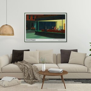 May include: A framed print of Edward Hopper's painting "Nighthawks" depicting a diner at night with three customers and a server. The painting is in a white frame with a black mat and is hanging on a white wall above a beige sofa.
