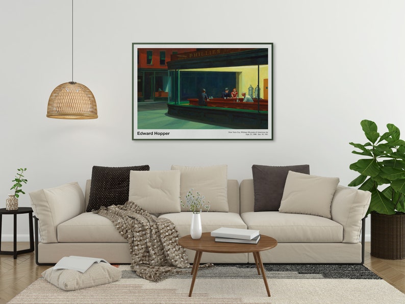 May include: A framed print of Edward Hopper's painting "Nighthawks" depicting a diner at night with three customers and a server. The painting is in a white frame with a black mat and is hanging on a white wall above a beige sofa.