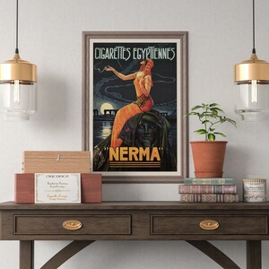 May include: A vintage poster advertising "Cigarettes Egyptiennes Nerma" with a woman in a flowing orange dress smoking a cigarette. The poster is framed and hanging on a wall above a wooden table with a plant, books, and a candle.