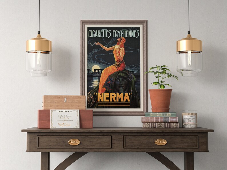 May include: A vintage poster advertising "Cigarettes Egyptiennes Nerma" with a woman in a flowing orange dress smoking a cigarette. The poster is framed and hanging on a wall above a wooden table with a plant, books, and a candle.