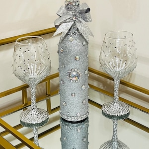 May include: A silver glitter wine bottle with a silver glitter bow and two silver glitter wine glasses on a gold tray.