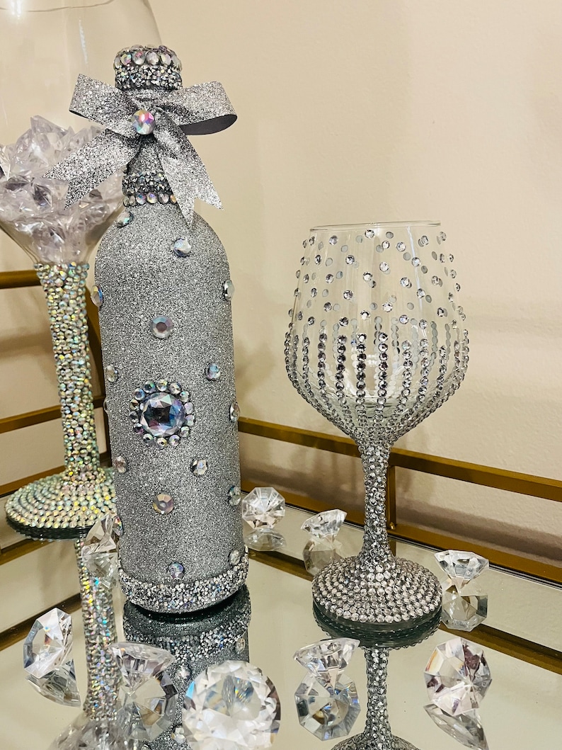 May include: A silver glitter wine bottle with a silver bow and a silver glitter wine glass. Both the bottle and glass are decorated with rhinestones.