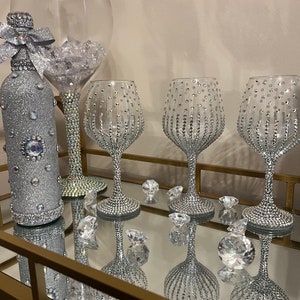 May include: A set of four silver-colored wine glasses and a bottle, all decorated with rhinestones. The glasses are on a gold-colored tray with a mirrored surface.