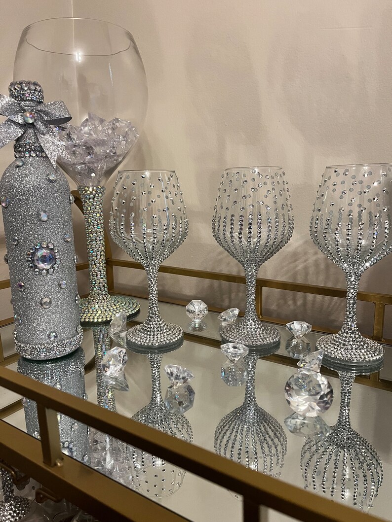 May include: A set of four silver-colored wine glasses and a bottle, all decorated with rhinestones. The glasses are on a gold-colored tray with a mirrored surface.
