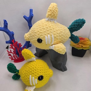 May include: Two handmade crochet fish toys featuring yellow bodies, dark green tails, and white gill details. Each fish has black plastic safety eyes. The larger fish is positioned on a gray stand, while the smaller fish rests below it.