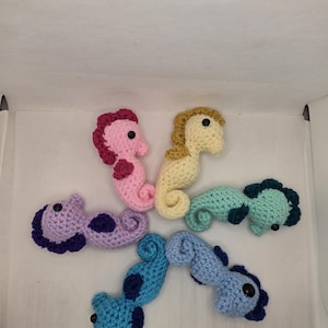 May include: Five crocheted seahorse toys arranged in a circular pattern. The seahorses are colored pink, pale yellow, mint green, light blue, and lavender, each featuring dark blue or purple fins, crests, and small black bead eyes.