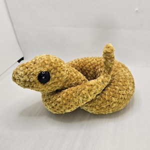 May include: A plush, golden-yellow snake toy with a textured, velvety appearance. The snake is coiled, with a raised tail and a black button eye. The toy is designed to resemble a snake.