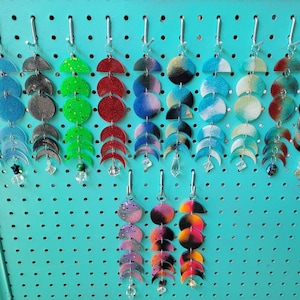 May include: A display of 12 pairs of dangle earrings featuring a crescent moon design. The earrings are made of resin and have a variety of colors and patterns, including glitter, tie-dye, and solid colors.