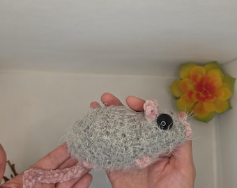 May include: A small, fuzzy, gray crocheted mouse toy with pink accents on the ears, nose, and tail. The mouse features a single visible black plastic bead eye and a textured, yarn-based body construction with a long, thin, braided pink tail.