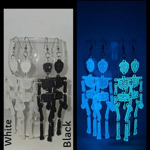 May include: Two pairs of skeleton earrings, one pair is white and the other is black. The earrings are shown in a glass dish. The earrings glow in the dark, and the glow is shown in the right side of the image.