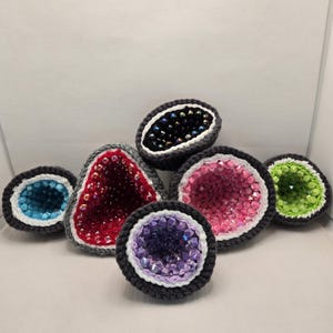 May include: A collection of crocheted geode-shaped objects in various colors. Each geode features a dark gray crocheted border with a white accent, and is filled with colorful beads. The geodes are in shades of blue, red, pink, purple, and green.