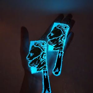 May include: A pair of glow-in-the-dark earrings shaped like butcher knives with a human face on the blade. The earrings are a light blue color and are glowing in the dark.