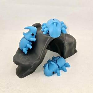 May include: Three blue plastic rhinoceros beetle figurines on a gray 3D printed rock. The beetles have black eyes and are posed in different positions.
