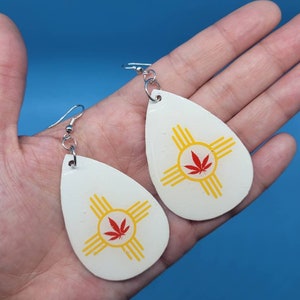 May include: A pair of white teardrop-shaped earrings with a red marijuana leaf and yellow Zia symbol on each earring.