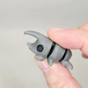 May include: A small, gray, 3D printed figurine of a beetle with a black eye. The beetle has a long, pointed horn and segmented legs.