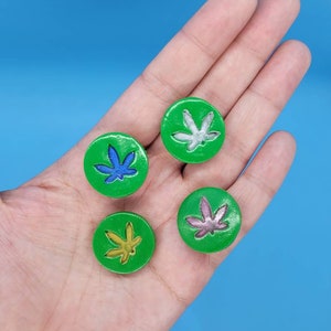 May include: Four green round buttons with a marijuana leaf design in different colors. The leaves are white, blue, yellow, and pink.