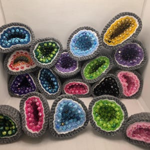 May include: A collection of crocheted geode-shaped ornaments featuring gray yarn borders, white inner rings, and centers filled with various colored beads including blue, green, yellow, purple, pink, and black, arranged in a cluster.