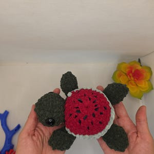 May include: A crocheted turtle plush toy featuring a dark green head and flippers, with a shell designed to resemble a watermelon slice, showcasing a vibrant red center with black seed-like accents and a light green border around the edge.