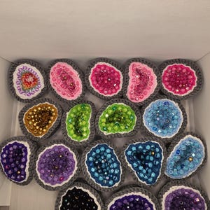 May include: A collection of fifteen crocheted geode-shaped ornaments, each featuring a grey yarn border, a white inner ring, and a center filled with various colored beads including pink, blue, green, purple, gold, and black, arranged in a box.