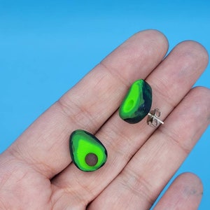 May include: A pair of avocado-shaped stud earrings. Each earring features a green and dark green outer layer with a bright green center and a brown seed. The earrings are held in a hand against a blue background.