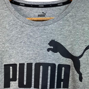 Peut inclure: Un t-shirt gris avec un logo Puma noir et une silhouette de Puma noire sautant.