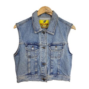 Op de afbeelding: Een lichtblauw denim vest met een geel label waarop "SQUAW" staat met een afbeelding van een schorpioen. Het vest heeft twee voorzakken en een knoopsluiting.