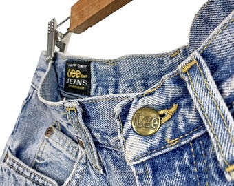 May include: Light blue denim jeans with a yellow stitched waistband and a brass button with the Lee logo. The label reads "Lee Jeans" in black text on a black background.