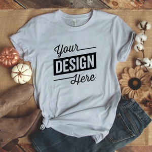 Könnte beinhalten: Ein hellgraues T-Shirt mit dem Text "Your Design Here" in Schwarz bedruckt. Das Shirt liegt auf einer Jutestoff-Oberfläche mit einem Paar braunen Stiefeln, Jeans und Herbstdeko.