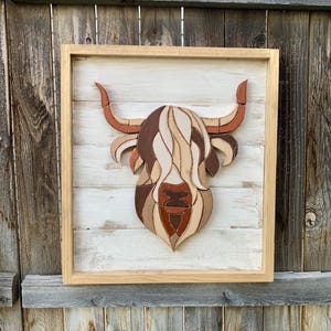 May include: Wooden wall art featuring a Highland cow head. The artwork is framed in a light wood frame and mounted on a white-washed wooden background. The cow's head is crafted from various shades of brown and tan wood pieces.