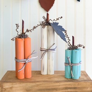 May include: Three wooden pumpkins, painted in orange, white, and teal, with twine accents and rustic wire stems. The pumpkins are arranged on a wooden shelf.
