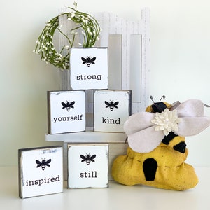 May include: Five small wooden signs with black bee silhouettes and inspirational words: strong, yourself, kind, inspired, still. A yellow and black bee-shaped stuffed toy sits in front of the signs.