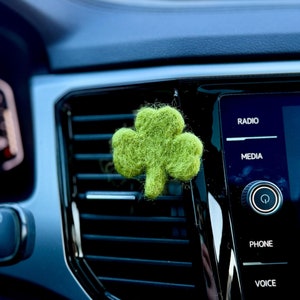May include: A green felt shamrock hanging from the air vent of a car's dashboard. The dashboard has buttons labeled "Radio", "Media", "Phone", and "Voice".