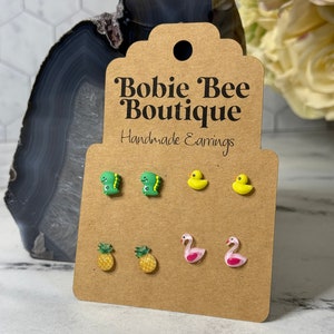 May include: A set of six pairs of handmade earrings featuring various summer-themed designs. The earrings include green dinosaurs, yellow ducks, yellow pineapples, and pink flamingos. The earrings are displayed on a brown card with the text "Bobie Bee Boutique Handmade Earrings".