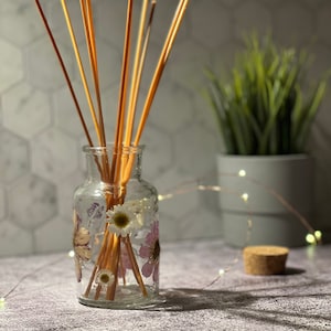 May include: A clear glass bottle with dried flowers and wooden diffuser sticks. The bottle has a floral design with pink and white flowers.