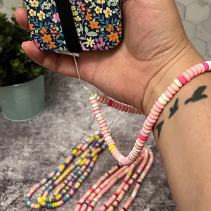 May include: A colorful beaded phone strap with pink, white, and yellow beads. The strap is attached to a phone case with a floral pattern.