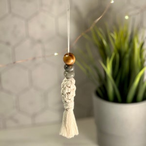May include: A white macrame tassel with a brown wooden bead and a gray stone bead. The tassel is hanging from a white string.