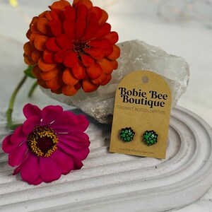 May include: A pair of green and black wooden earrings with a floral design. The earrings are handmade and are displayed on a card that reads "Bobbie Bee Boutique Handmade Wooden Earrings" and includes the website address "www.BobbieBeeBoutique.com".