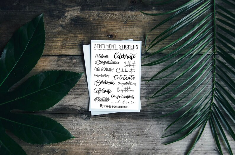 Puede incluir: Una tarjeta blanca con texto negro que dice "Sentiment Stickers" y presenta varias palabras escritas a mano, incluyendo "Celebrate" y "Congratulations". La tarjeta est&aacute; sobre una superficie de madera con hojas verdes.